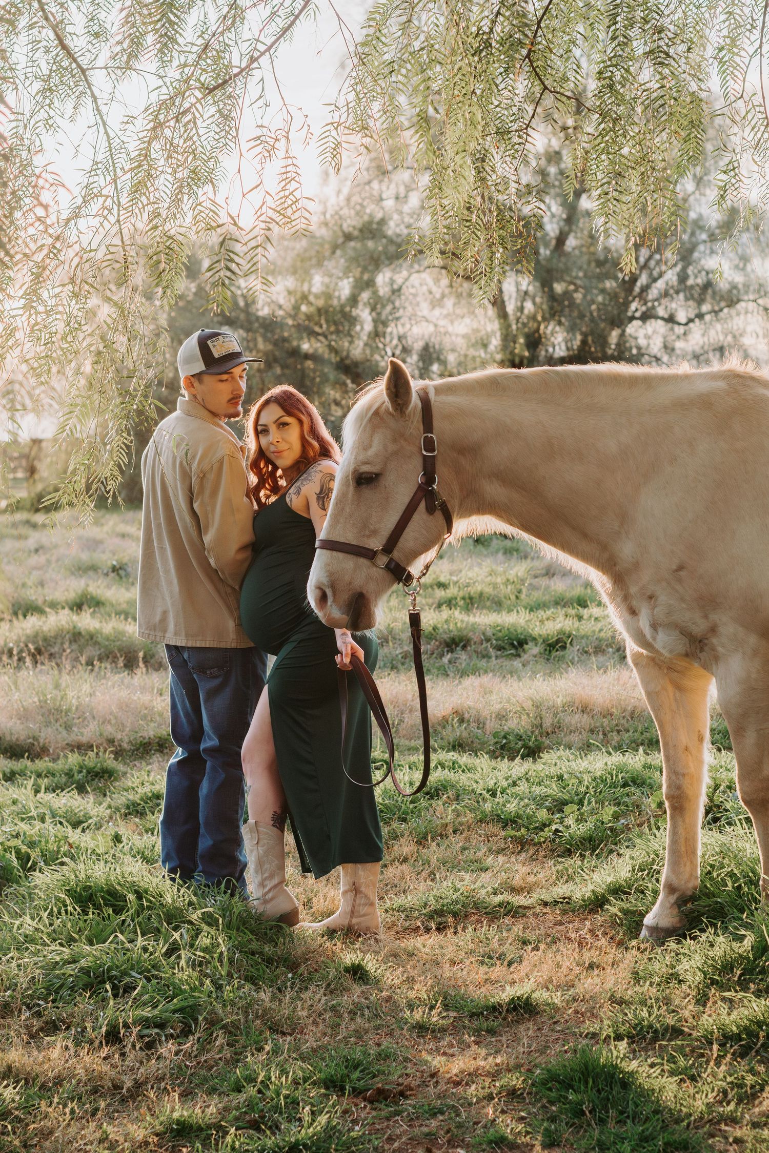 A couple stands together with a cream-colored horse under drooping willow branches in a sunlit meadow during a maternity photoshoot in Lodi.