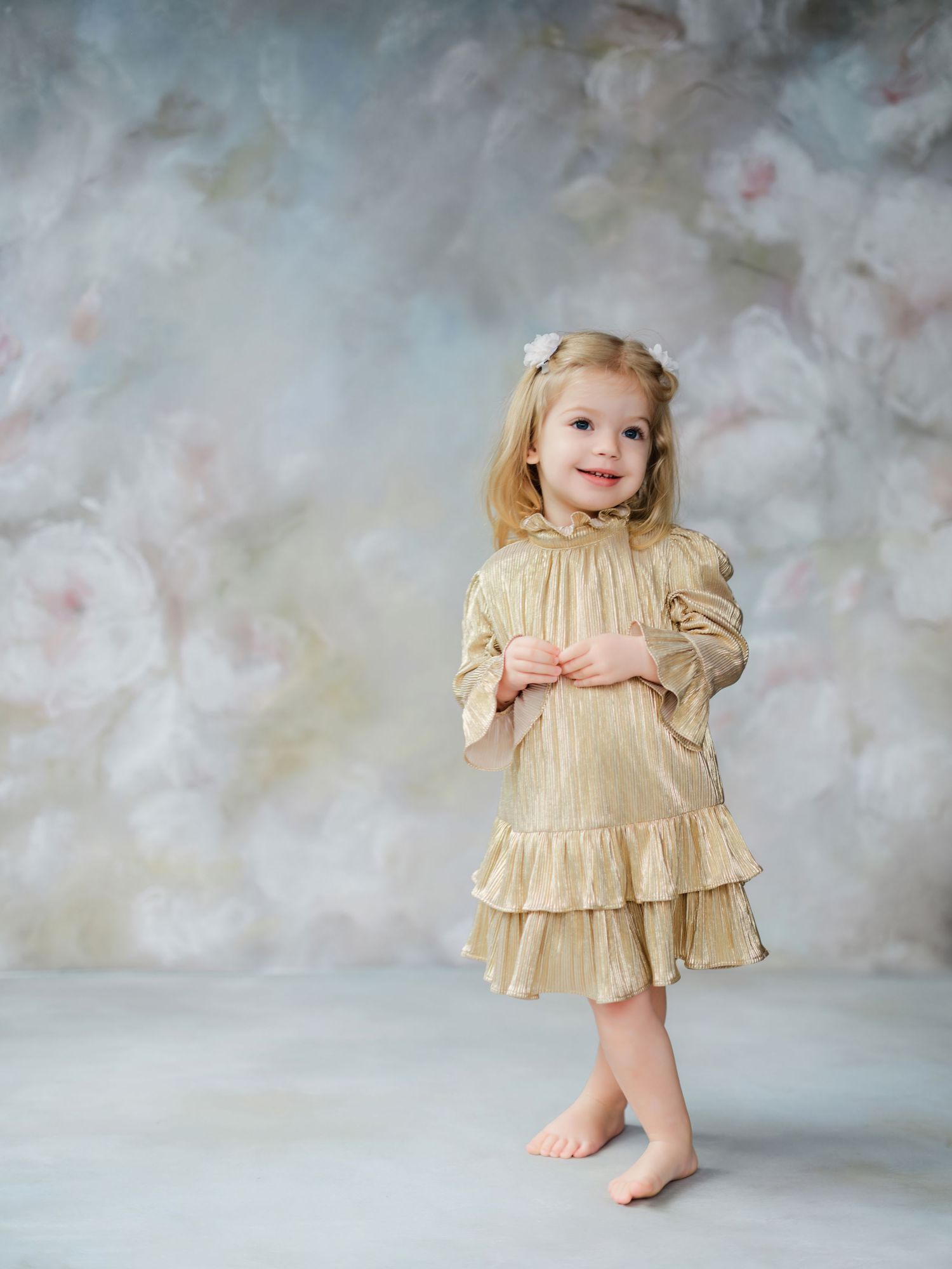 A small child stands barefoot wearing a vintage-style cream colored dress with ruffles against a soft cloudy backdrop.