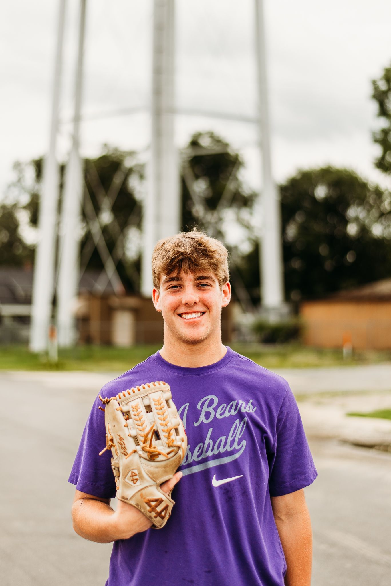 Baseball player wearing purple team shirt holds up tan leather glove against cloudy sky.