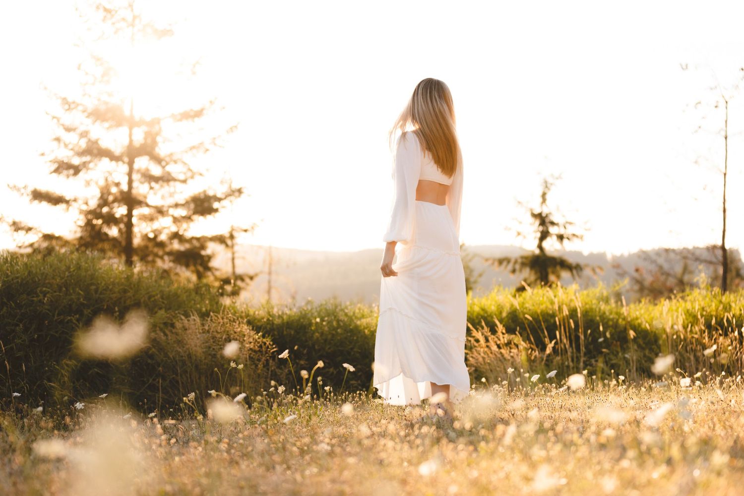 A person in a white dress stands in a sunlit meadow with wildflowers at golden hour.