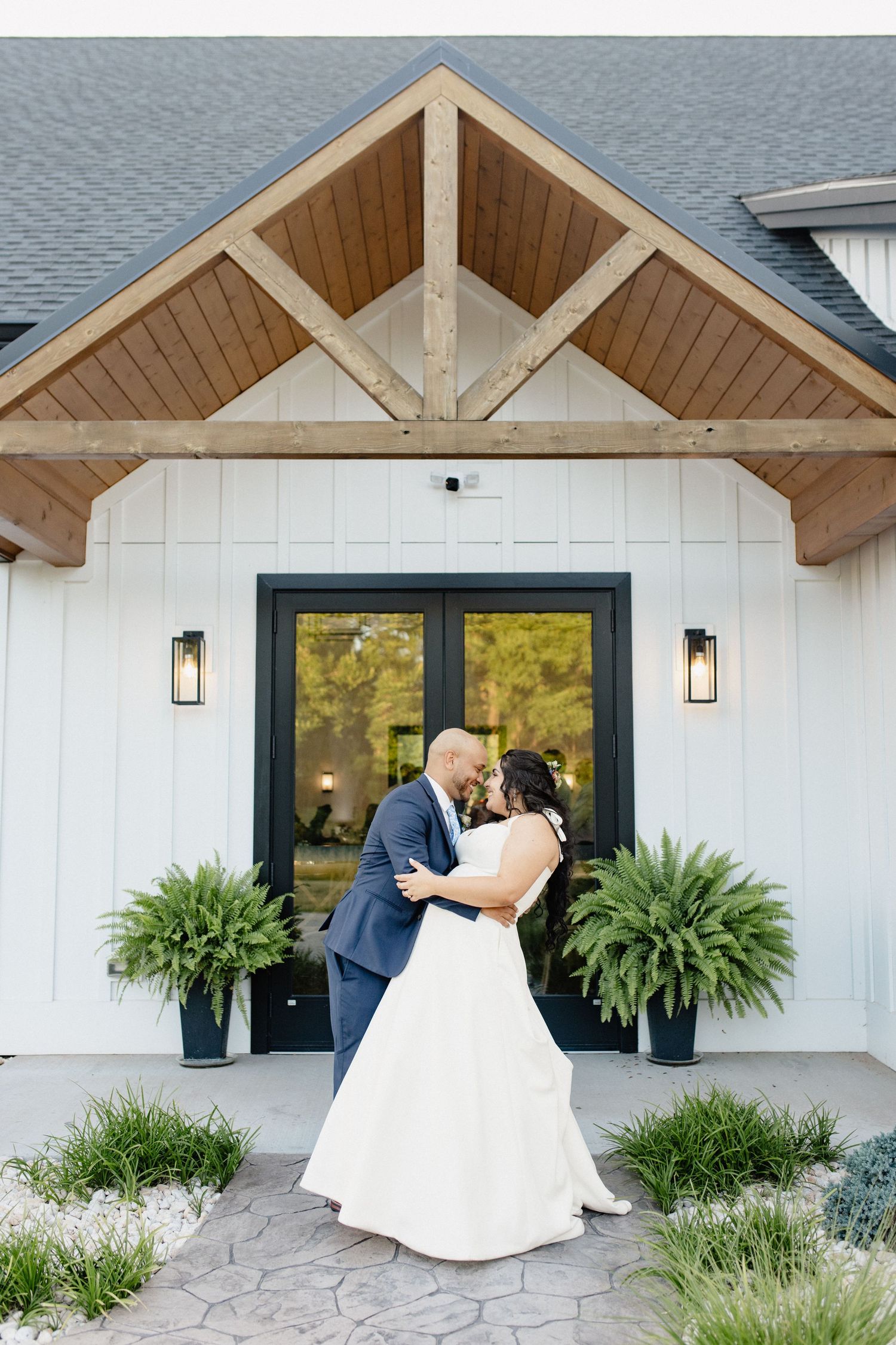A couple shares their first dance outside a modern farmhouse entrance with wooden beams.