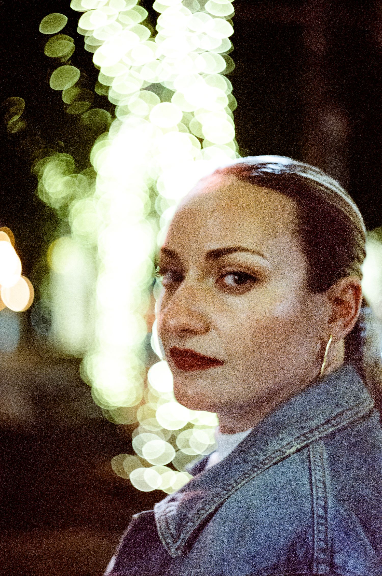 A woman in a denim jacket poses against a dark background with blurred bokeh lights creating a soft glow behind her.