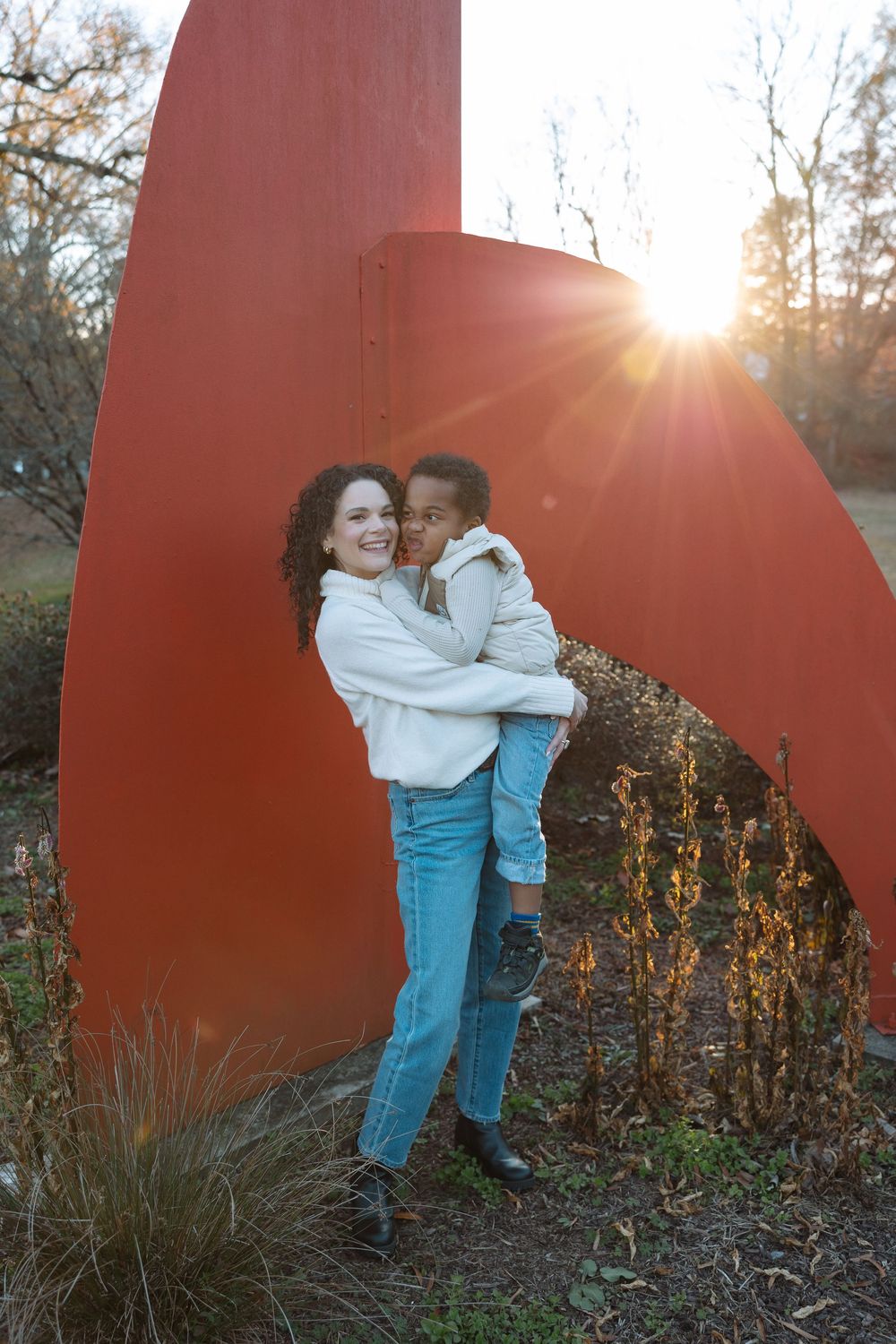 Two people embrace and smile while standing against a large red outdoor sculpture at sunset.