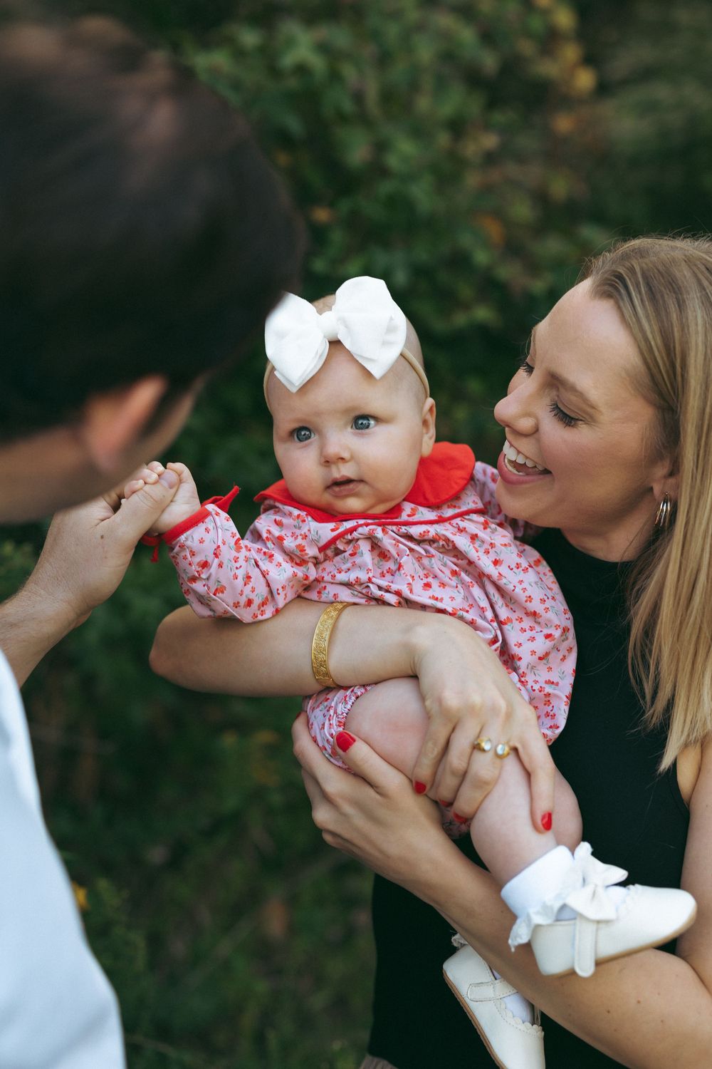 A baby in a red and white floral dress and bow being held and lovingly embraced outdoors.