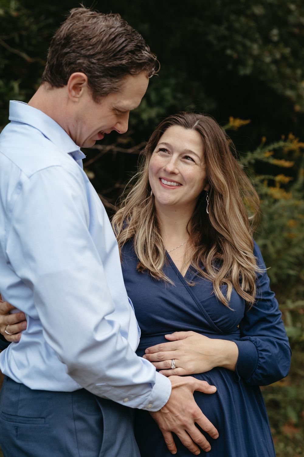 A couple shares an intimate moment outdoors, with the woman wearing a navy dress and the man in a light blue shirt.