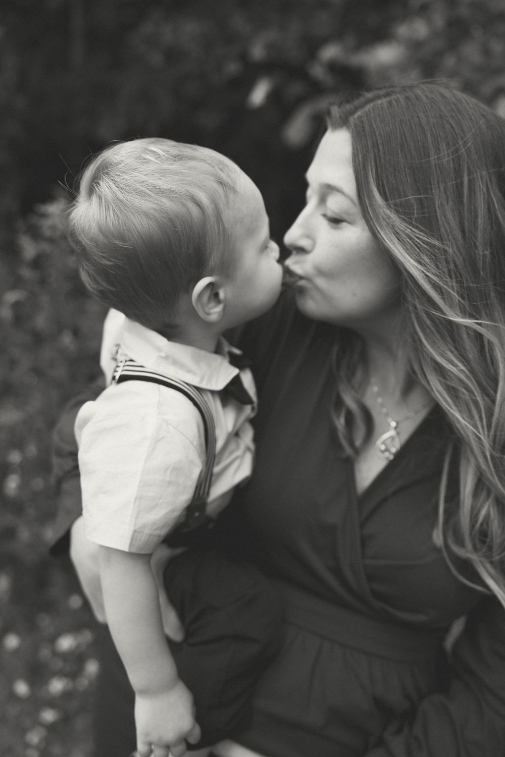 A tender black and white portrait of a mother kissing her young toddler against a blurred outdoor background.
