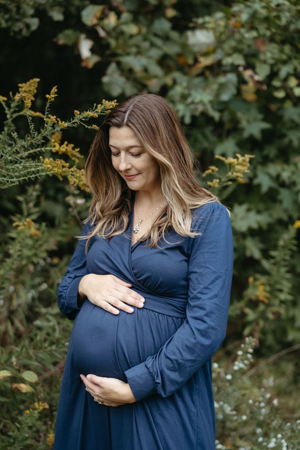 Expectant mother in a navy blue dress smiles while cradling her baby bump in a garden setting.
