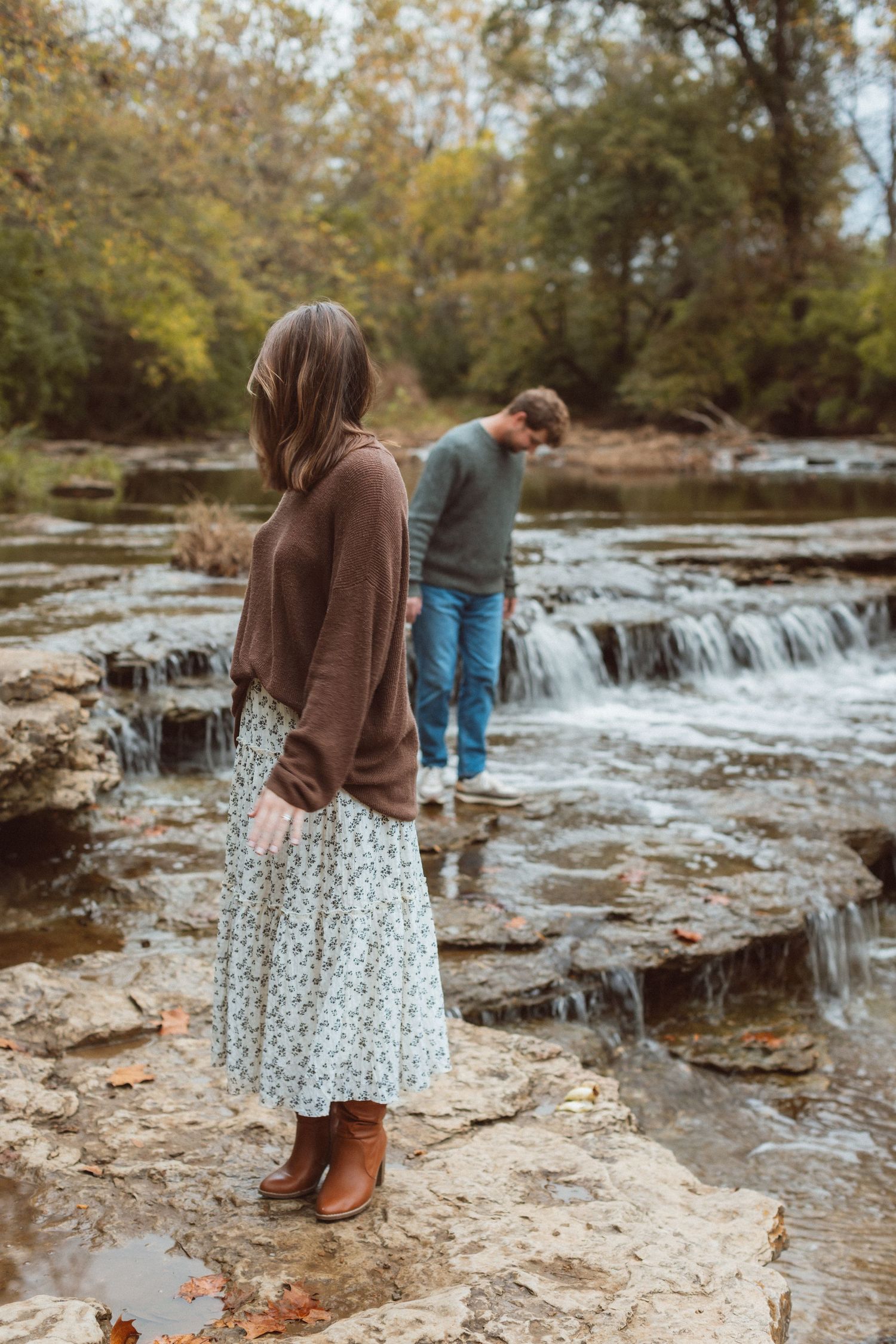 A woman looks back at a man as they explore a small waterfall in this scene from a fall engagement photo session in Kansas City.