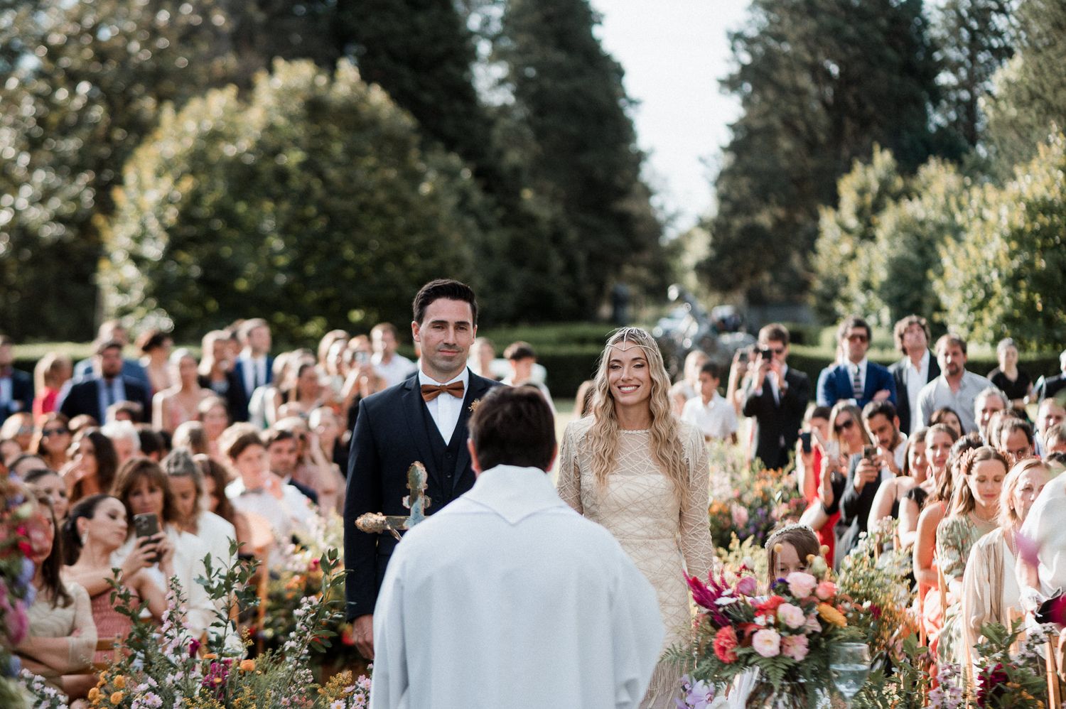 Wedding guests stand in rows outdoors during a ceremony surrounded by colorful summer flowers and greenery.