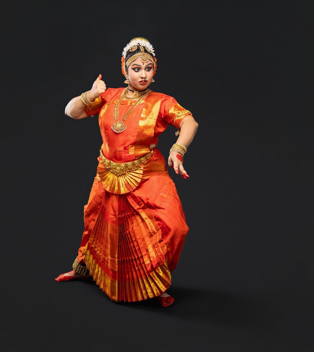 Classical Indian dancer performs in vibrant orange and red traditional costume with expressive hand gestures against dark backdrop.
