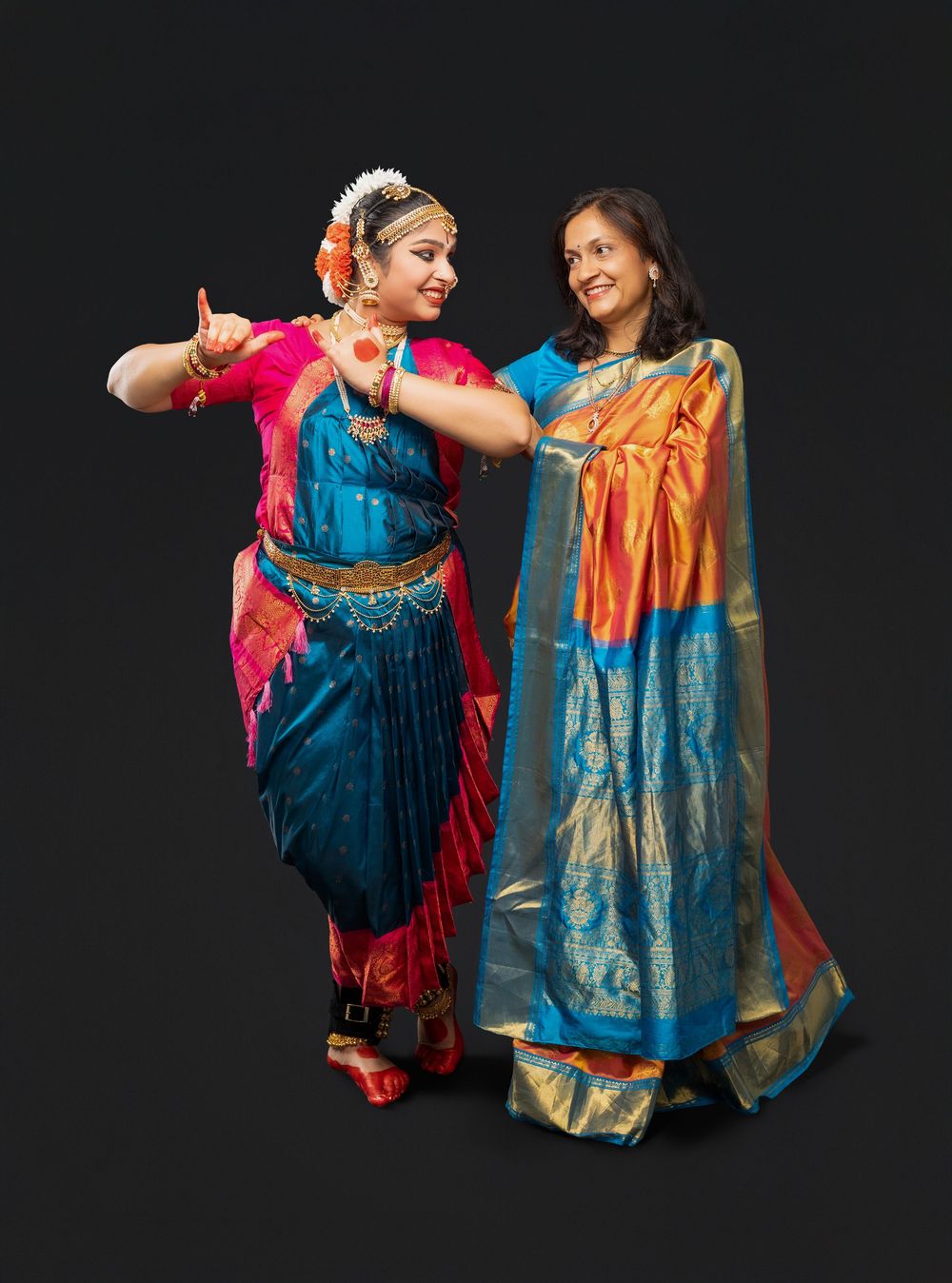 Two dancers in vibrant traditional Indian dance costumes perform expressive movements against a black background.