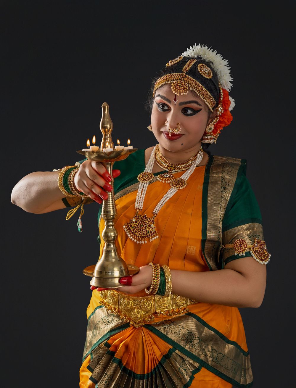 Traditional Indian classical dancer performs with oil lamp in orange and green costume against dark background.