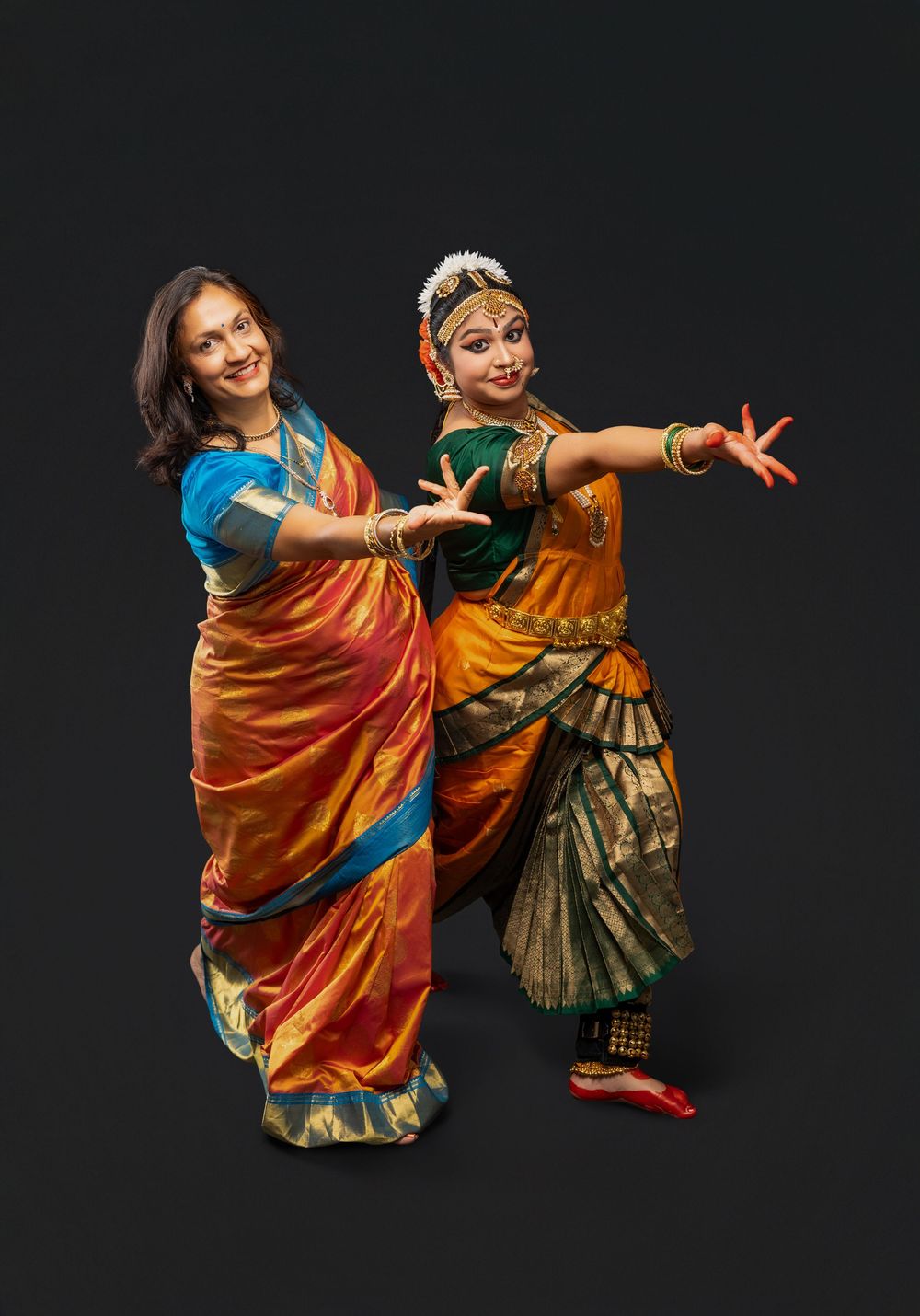 Two dancers in vibrant traditional Indian costumes perform a dynamic classical dance sequence against a dark background.
