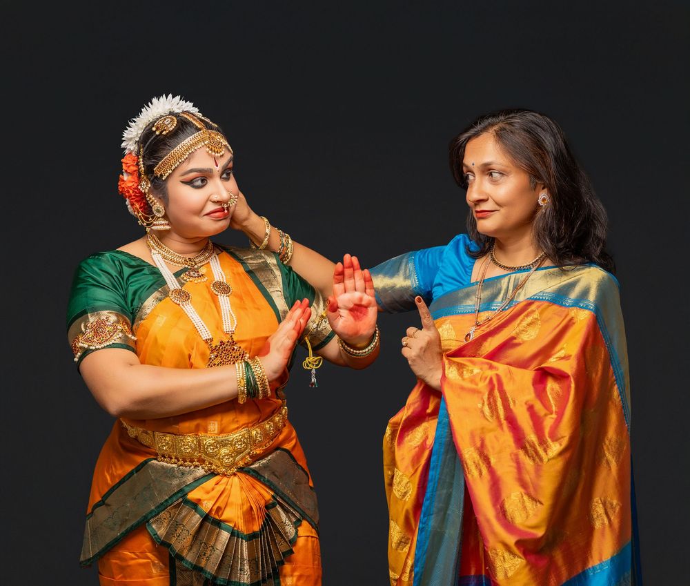 A group of dancers in vibrant orange and green traditional Indian saris perform a coordinated dance sequence against a dark background.