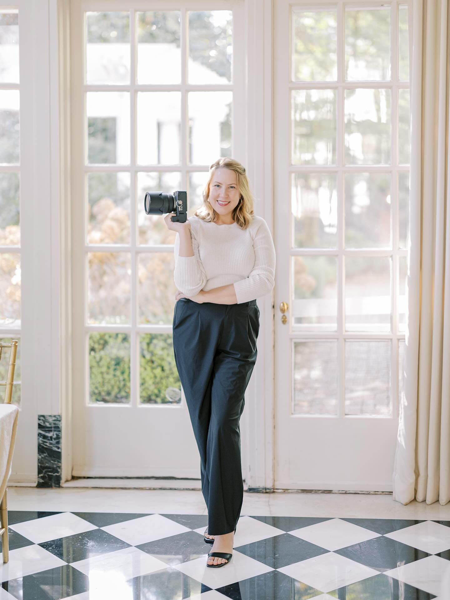 A person in a beige sweater and dark pants stands in a bright sunlit room with black and white checkered floors.