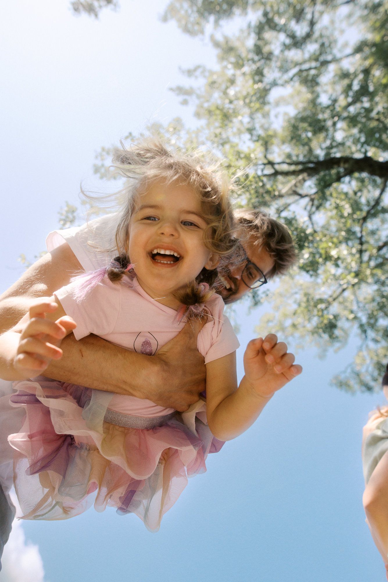 A child in a pink dress laughs joyfully while being lifted up against a bright blue sky with green trees.