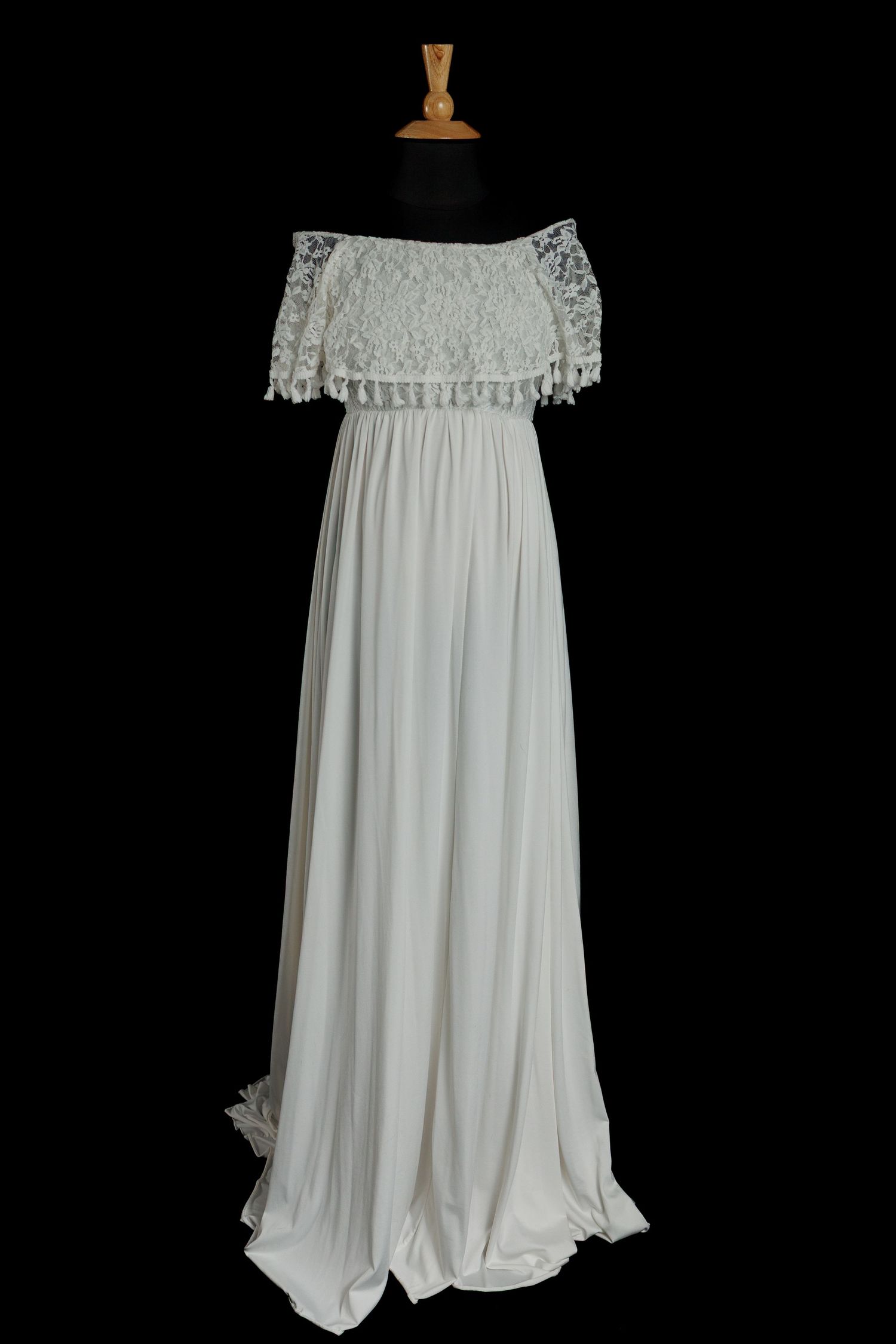 Ivory maternity gown with lace off-the-shoulder bodice, tassel-trim sleeves, empire waist, and flowing skirt on a dress form.