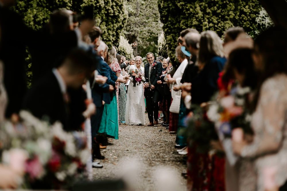 Wedding guests line a garden path while celebrating the newlyweds in an outdoor ceremony.