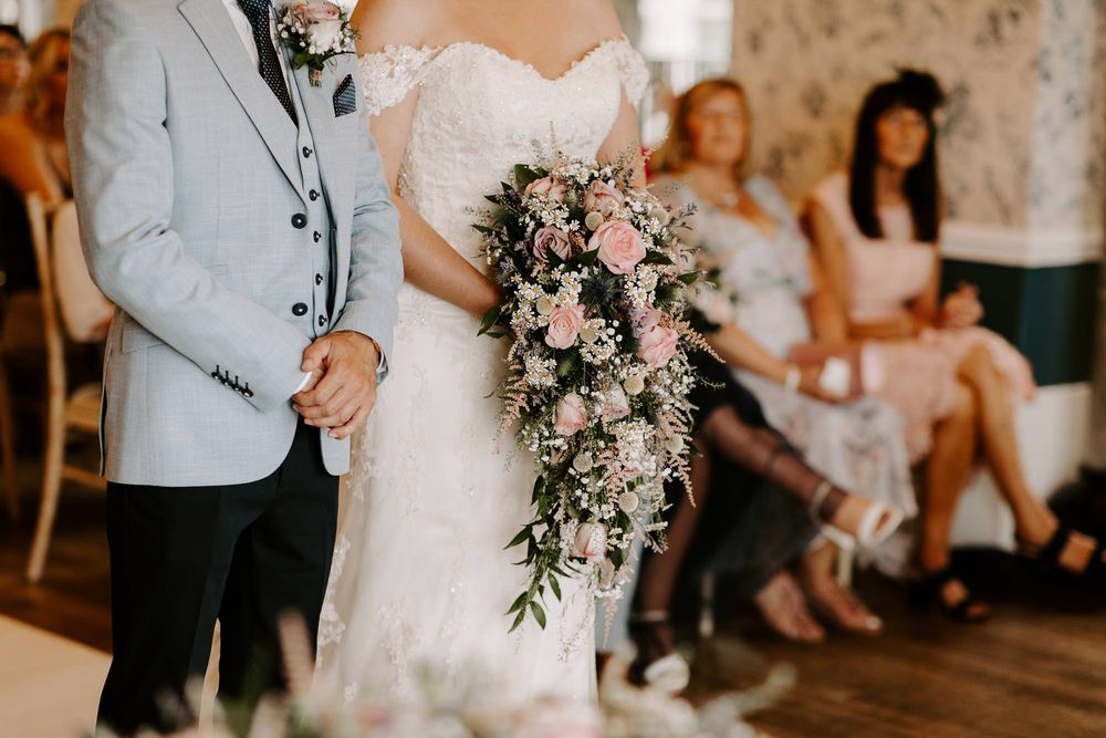A cascading bridal bouquet with pink roses and greenery is held during an indoor wedding ceremony.