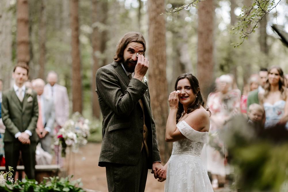 A romantic outdoor wedding ceremony in a wooded forest setting with guests watching the couple exchange vows.