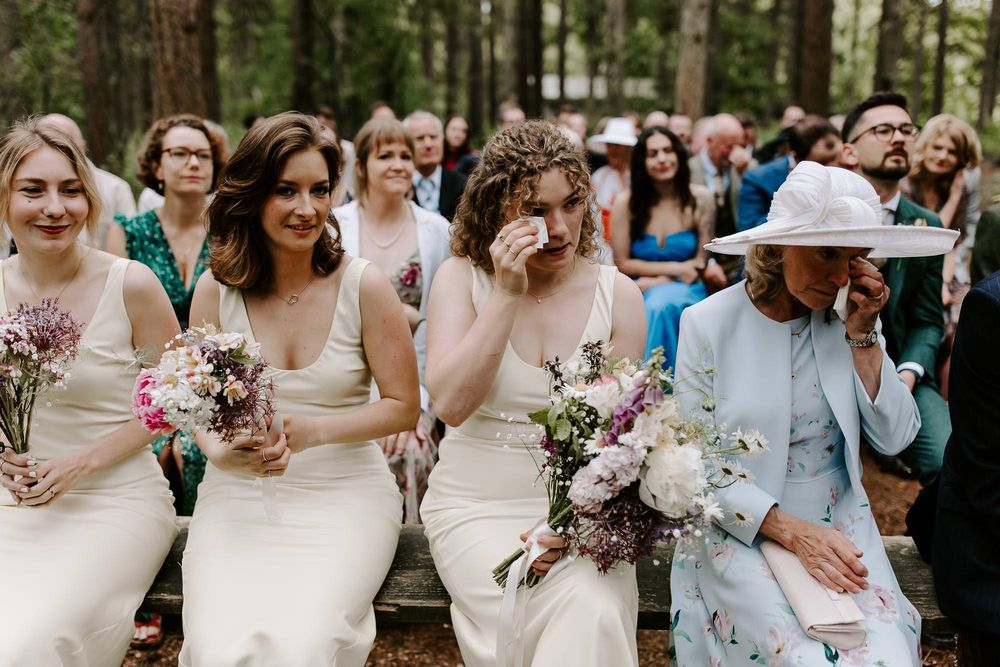 A wedding party stands together in a forest setting wearing light blue and cream colored attire with floral bouquets.