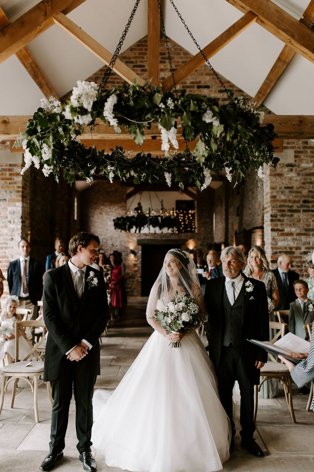 Wedding ceremony in a rustic barn venue with exposed wooden beams and brick walls decorated with greenery and string lights.