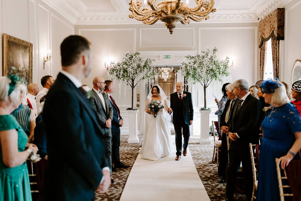 Wedding guests gather in an elegant white ballroom with ornate chandeliers and decorative molding during a reception.