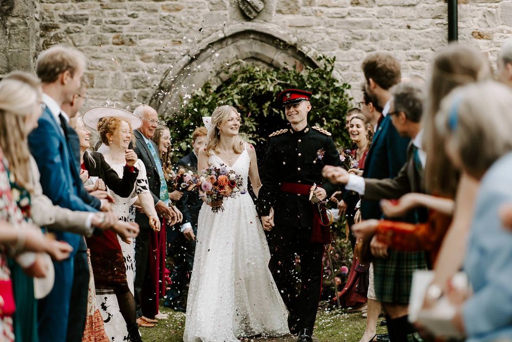 Wedding guests celebrate with confetti and bubbles during an outdoor ceremony at a rustic stone venue.
