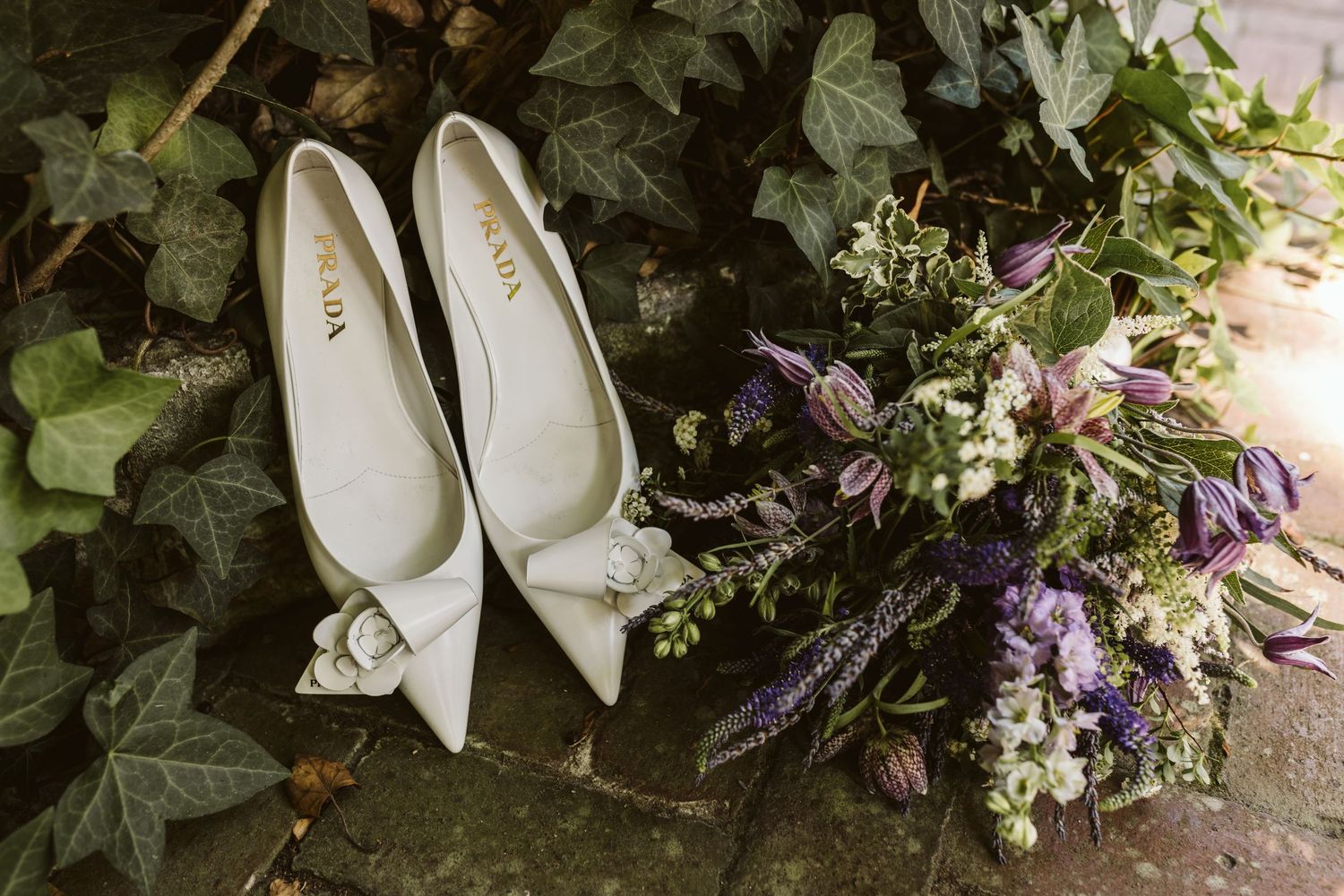 Elegant white bridal shoes surrounded by delicate purple and white floral arrangements and greenery.