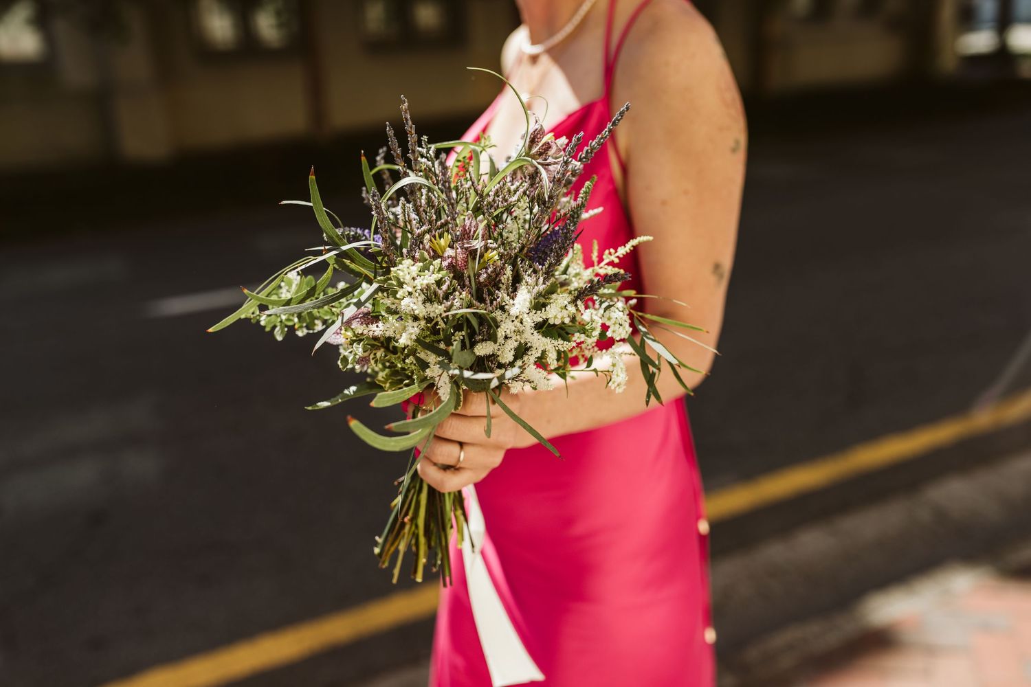 Someone in a vibrant pink dress holds a rustic wildflower and greenery bouquet against a dark background.