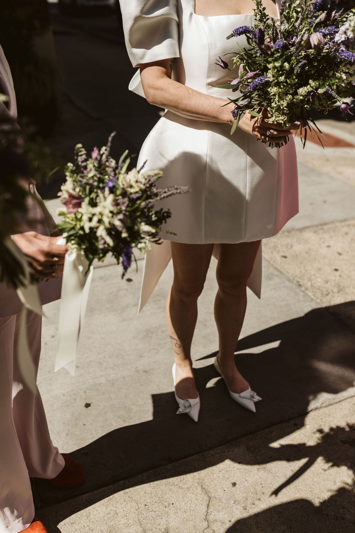 Wedding guests in white dresses walk along a sunlit pathway holding floral bouquets.