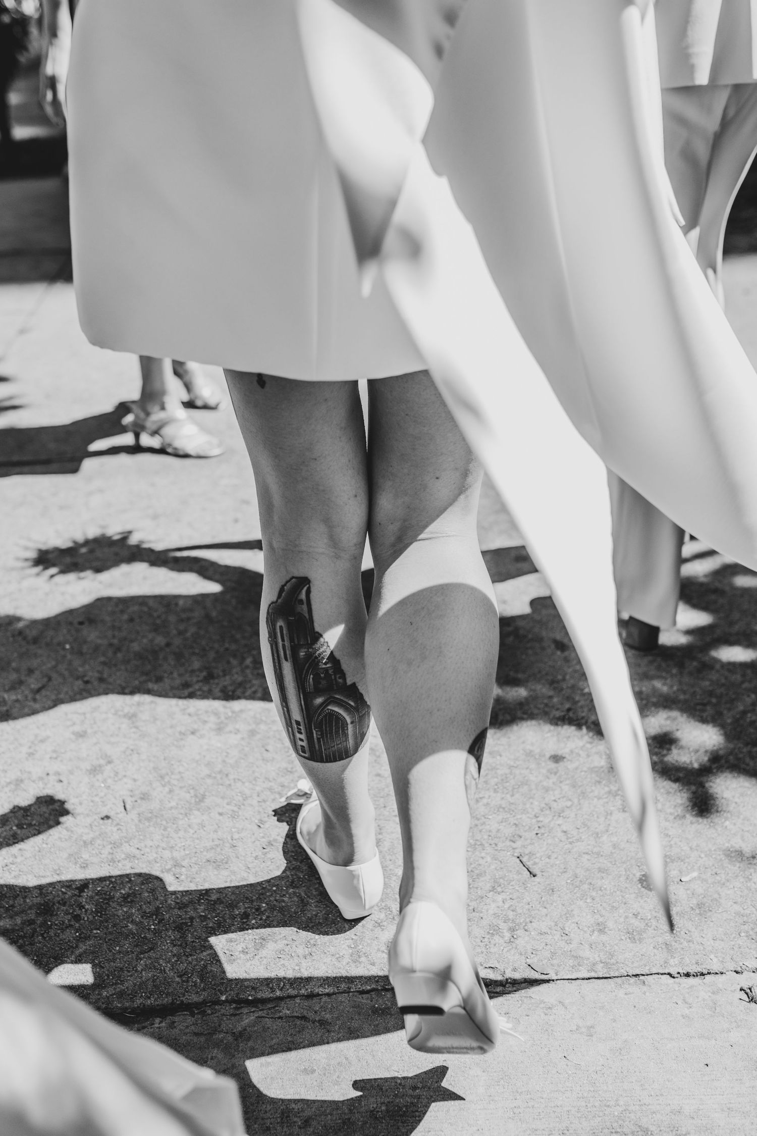 Black and white image showing legs in white shoes and skirts walking on a sunny outdoor surface.