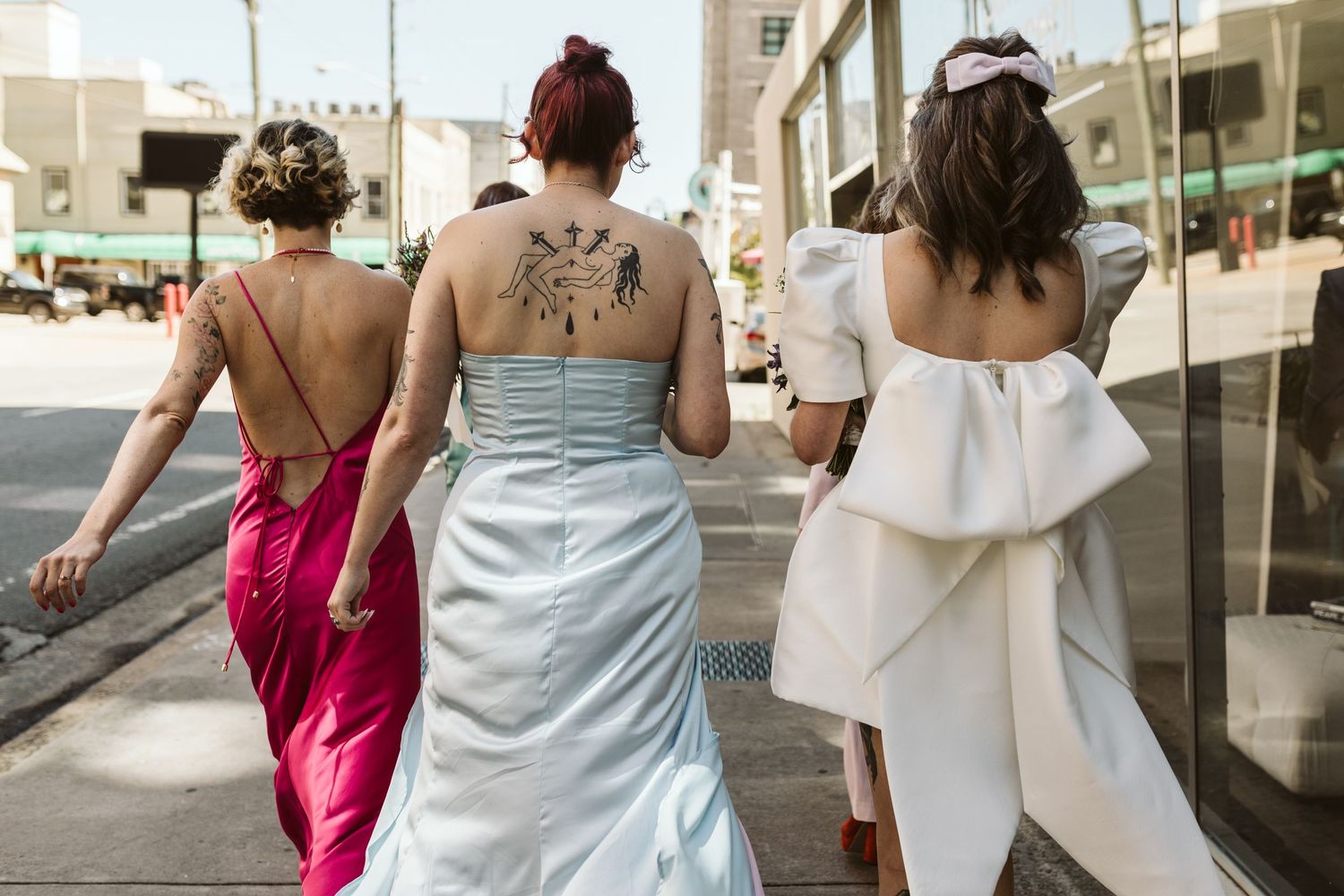 Three friends in summer outfits walk together down a city sidewalk while chatting and smiling.