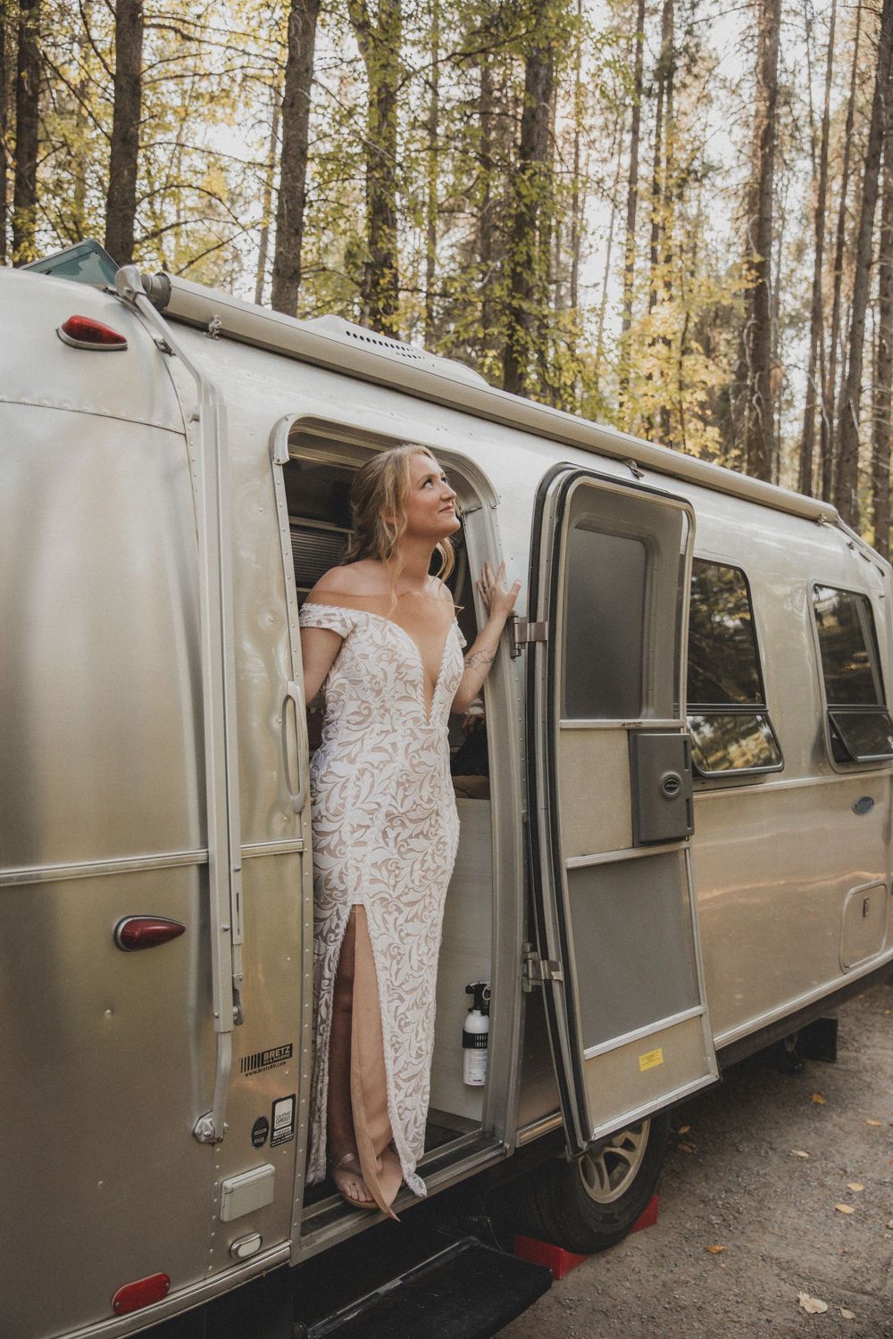 White lace dress detail shown through vintage Airstream trailer windows in forest setting.