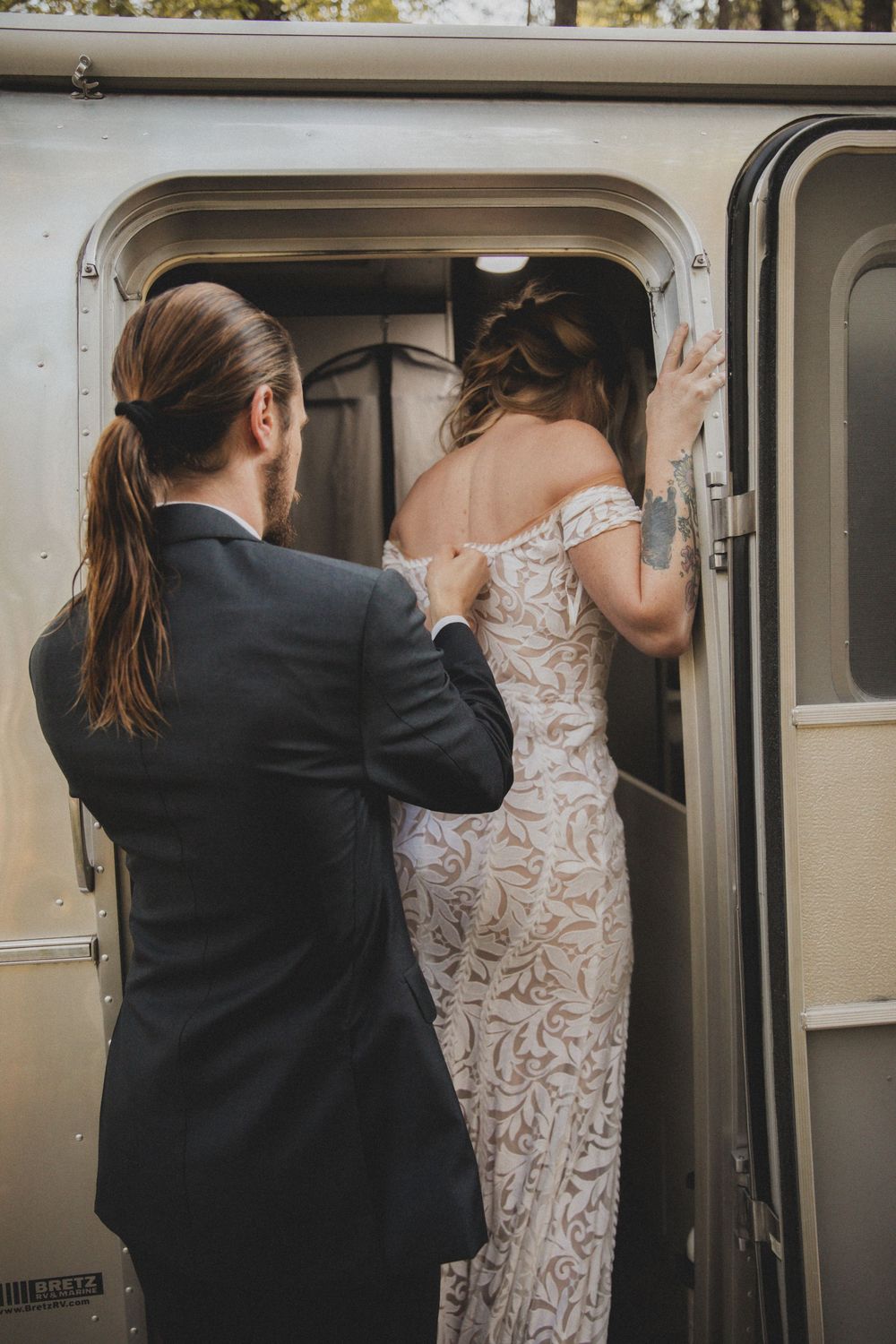 A couple in wedding attire entering a silver Airstream trailer in a wooded setting.