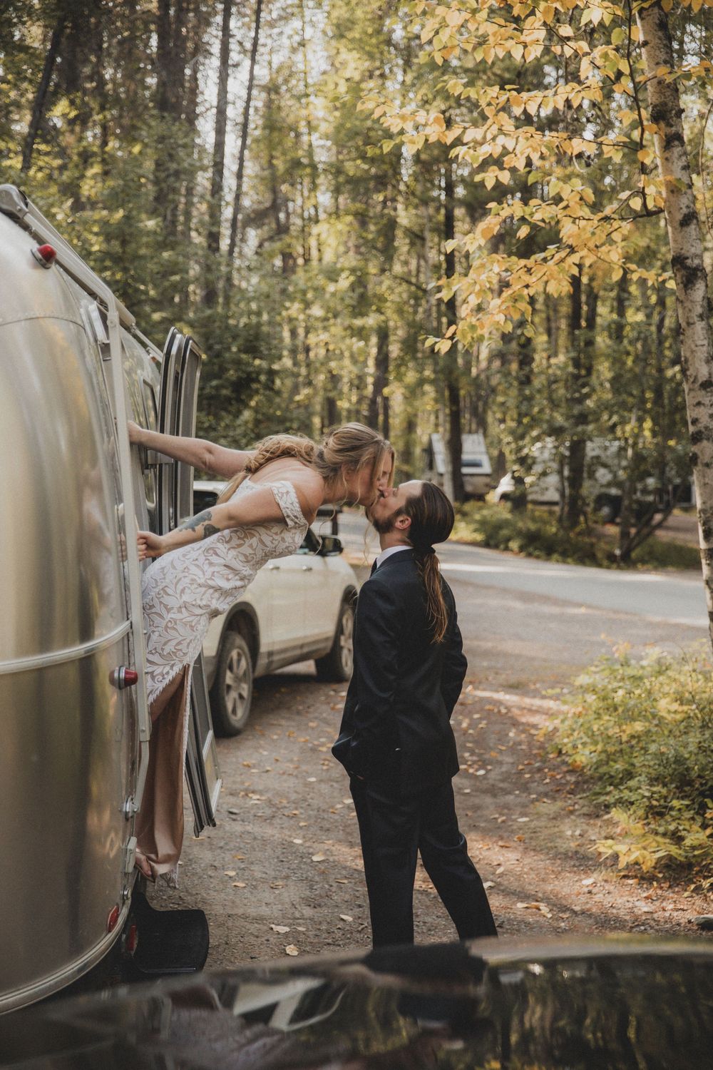 A couple poses playfully by a silver Airstream trailer in an autumn forest setting.