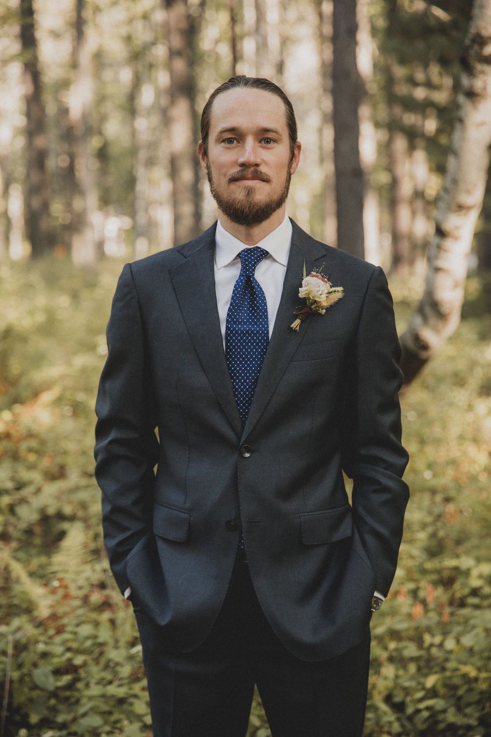 Three groomsmen in navy suits with blue ties and boutonnieres stand in a wooded forest setting.
