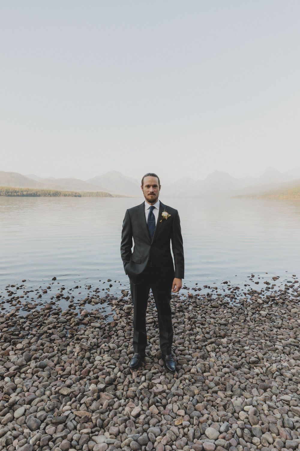 Well-dressed figures standing on rocky shoreline with mountains and lake in background.
