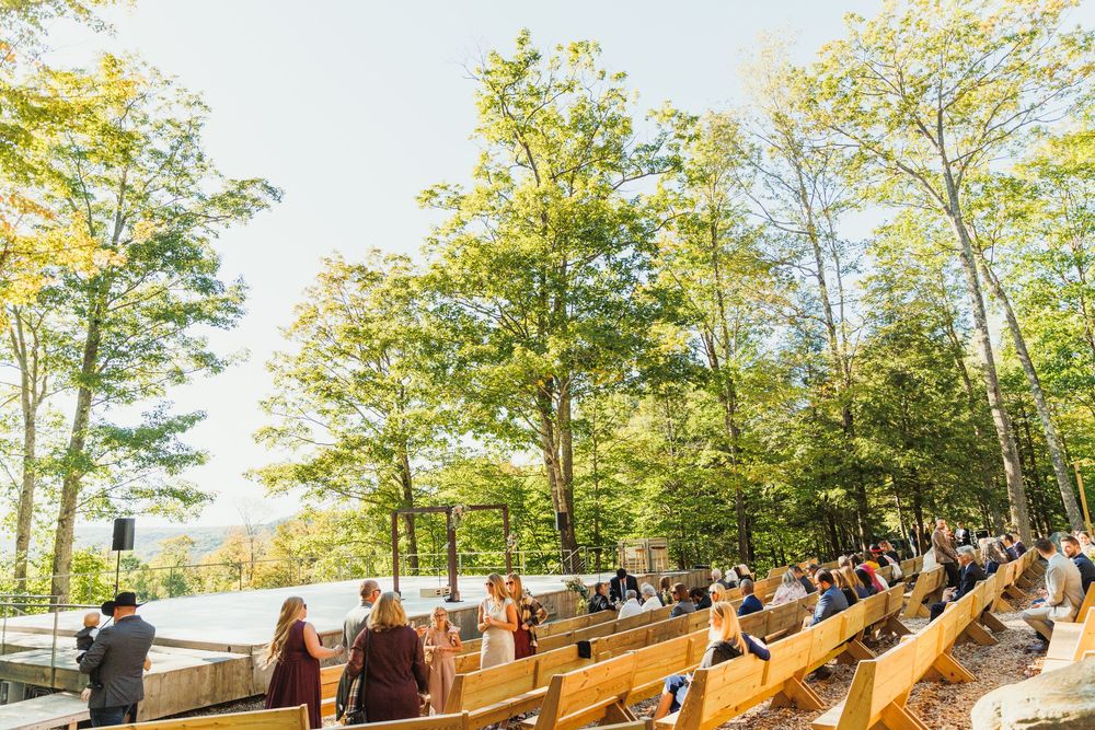 Panoramic view of outdoor wedding ceremony setup with wooden benches in wooded area.