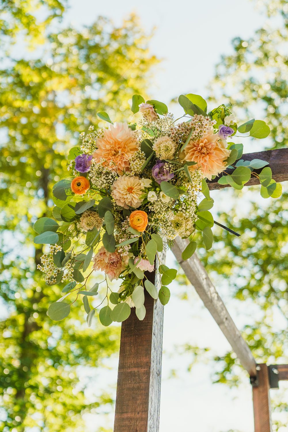 Peach and coral floral arrangement with greenery adorns a wooden wedding arch against sunlit trees.