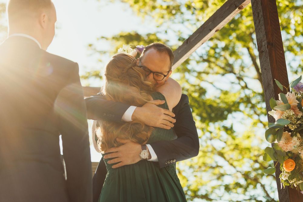 Emotional embrace during outdoor wedding ceremony under wooden pergola with floral decorations.