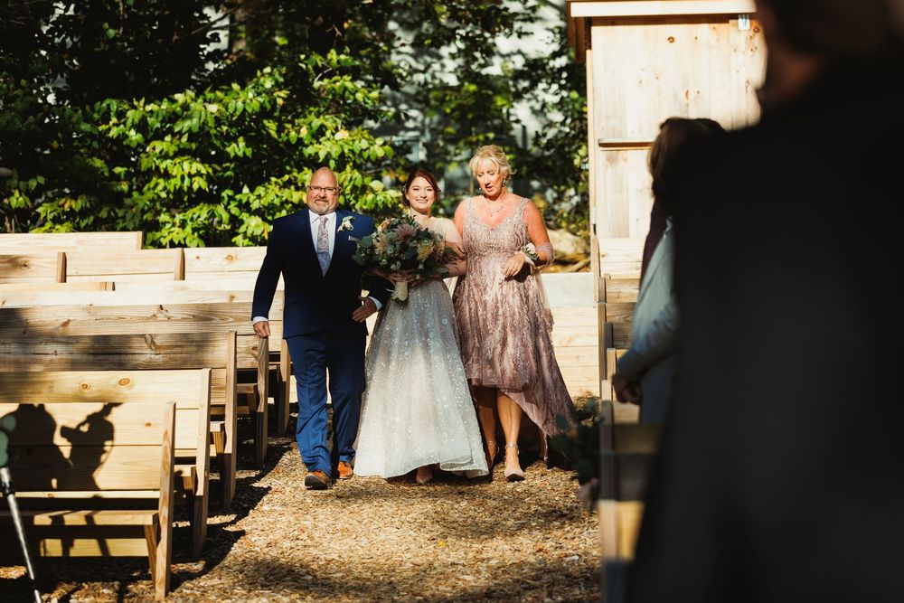 Wedding party walking down aisle during outdoor ceremony with rustic wooden backdrop.