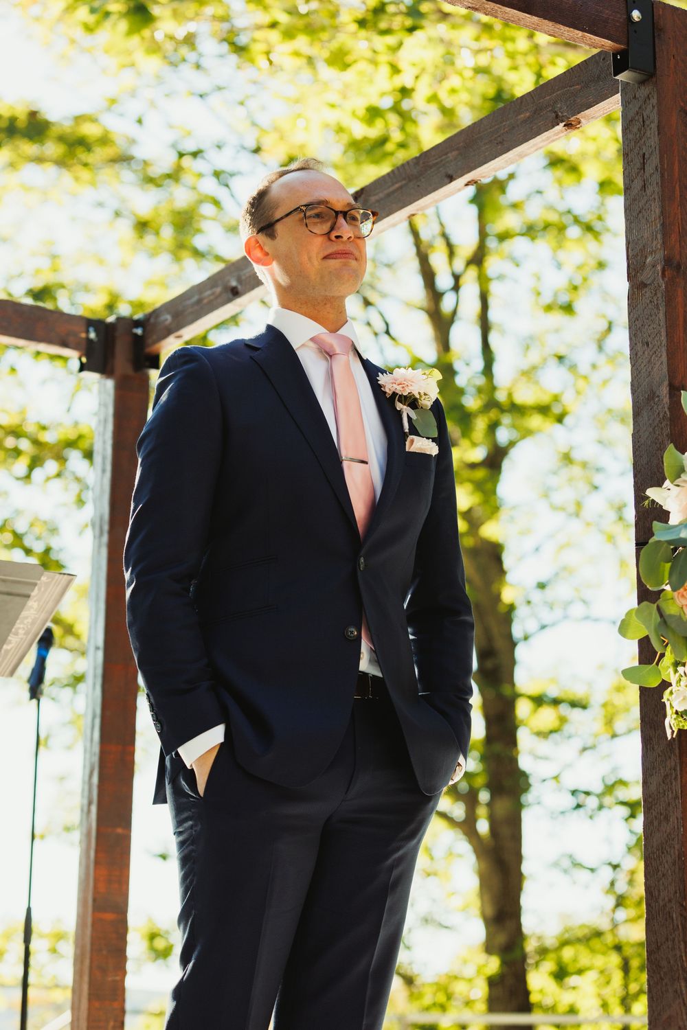 Groom in navy suit and pink tie stands under wooden pergola during outdoor ceremony.