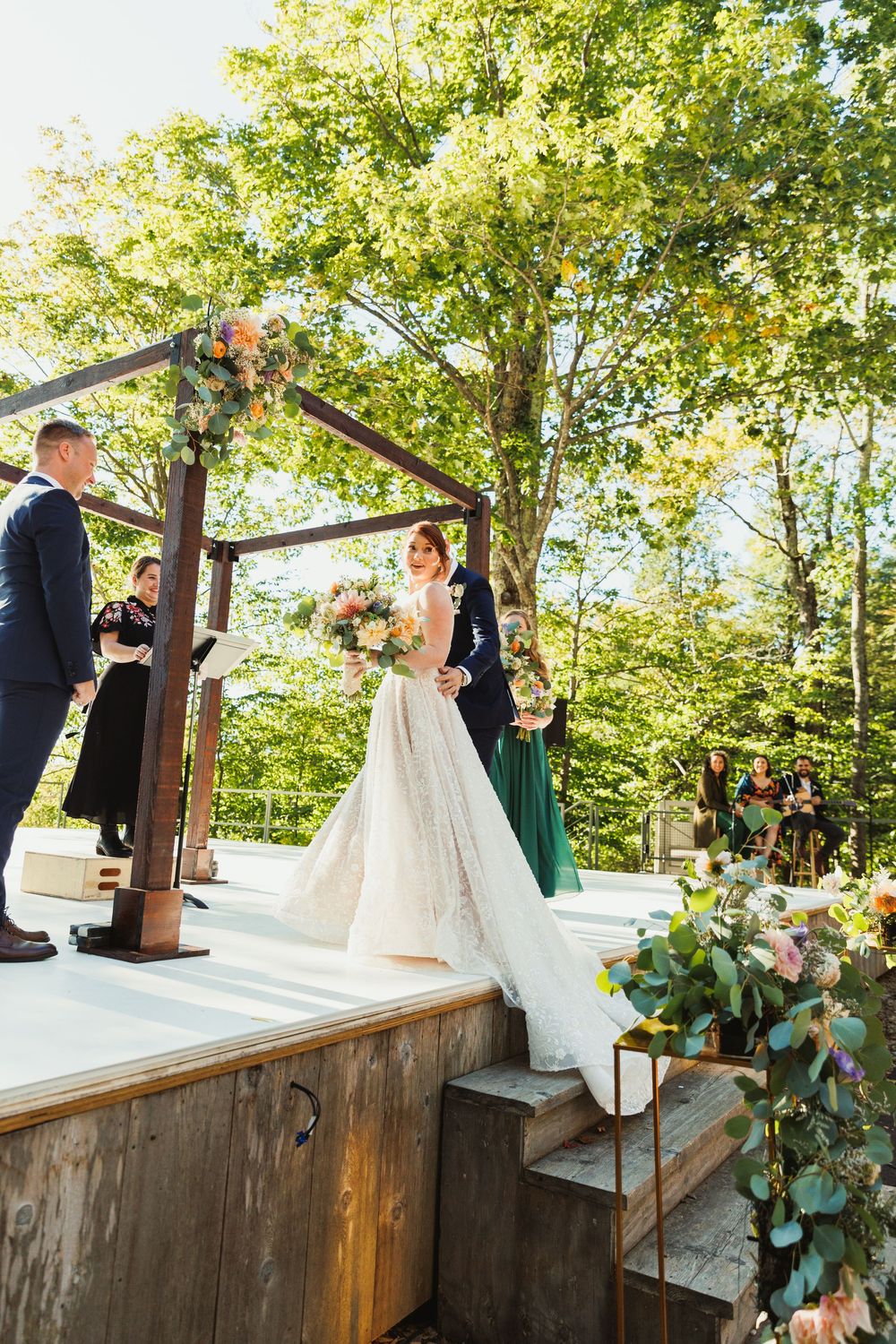 Wedding ceremony on elevated wooden platform with rustic pergola and floral arrangements.