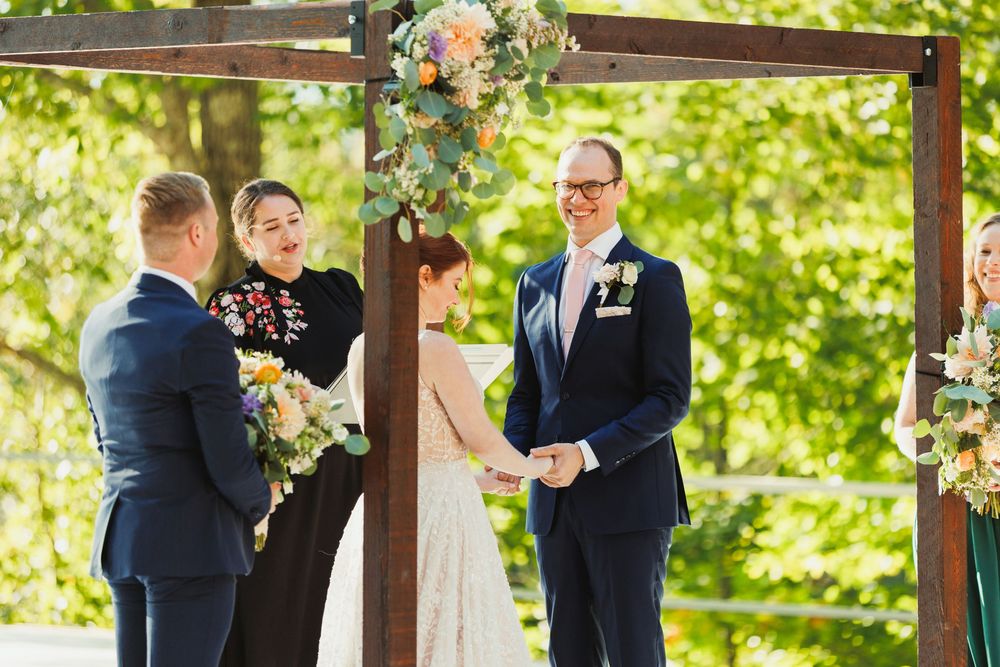 Outdoor wedding ceremony taking place under a wooden pergola decorated with floral arrangements on a sunny day.