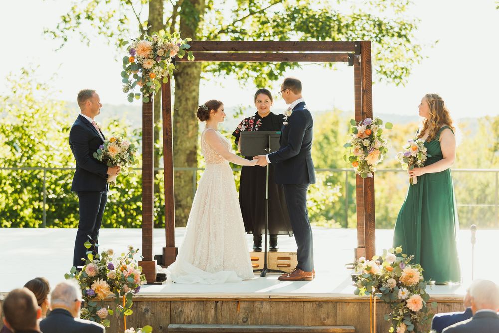 Wedding ceremony under a rustic wooden arbor with floral decorations and natural greenery in the background.
