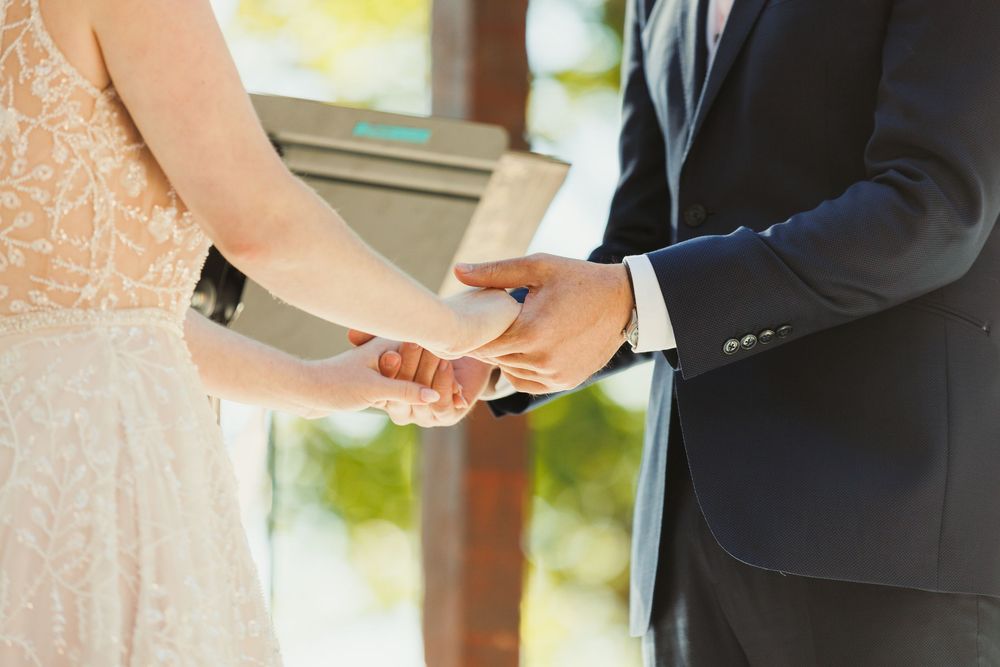 Close-up of hands during wedding ceremony exchange beneath a decorated wooden structure.