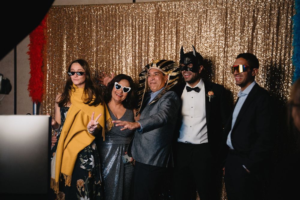 Five wedding guests posing with fun masks and sunglasses in front of a gold sequin photo booth backdrop.