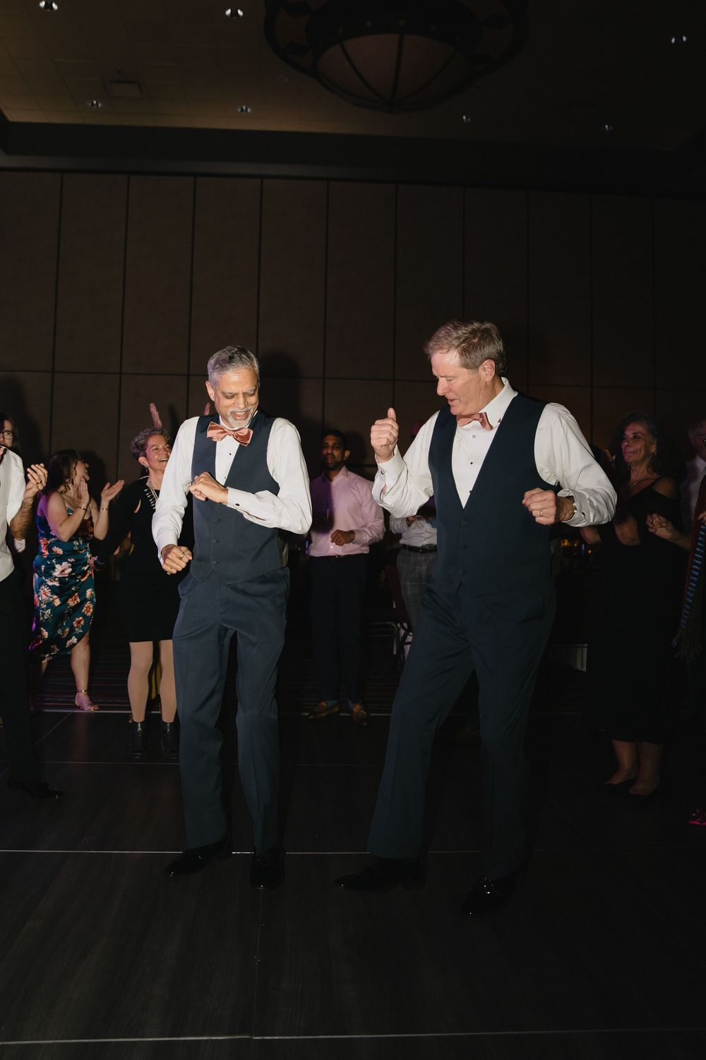Two older men in matching vests and bow ties dancing together at a wedding reception.