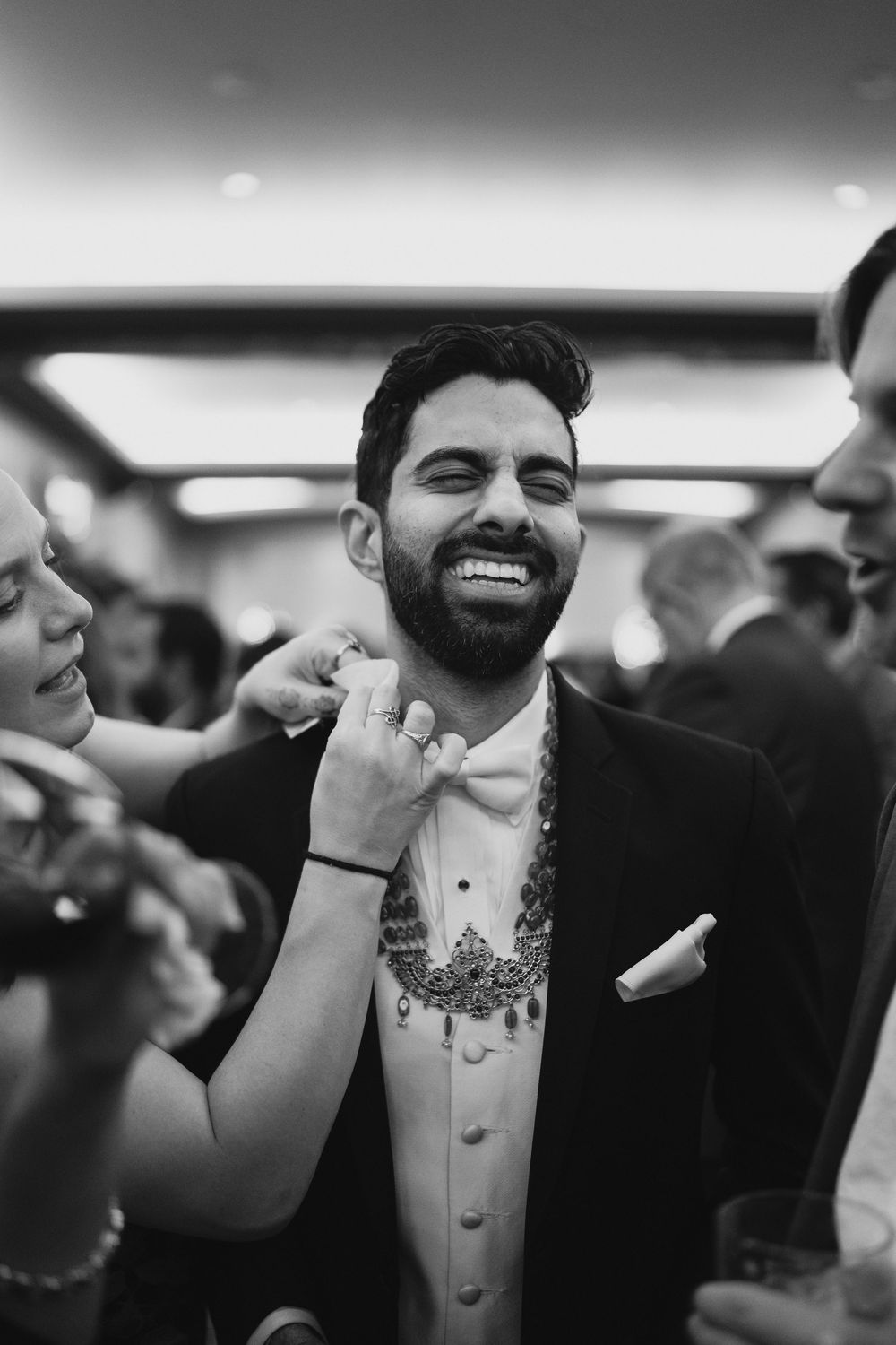 Black and white photo of smiling groom having necklace adjusted at wedding reception.